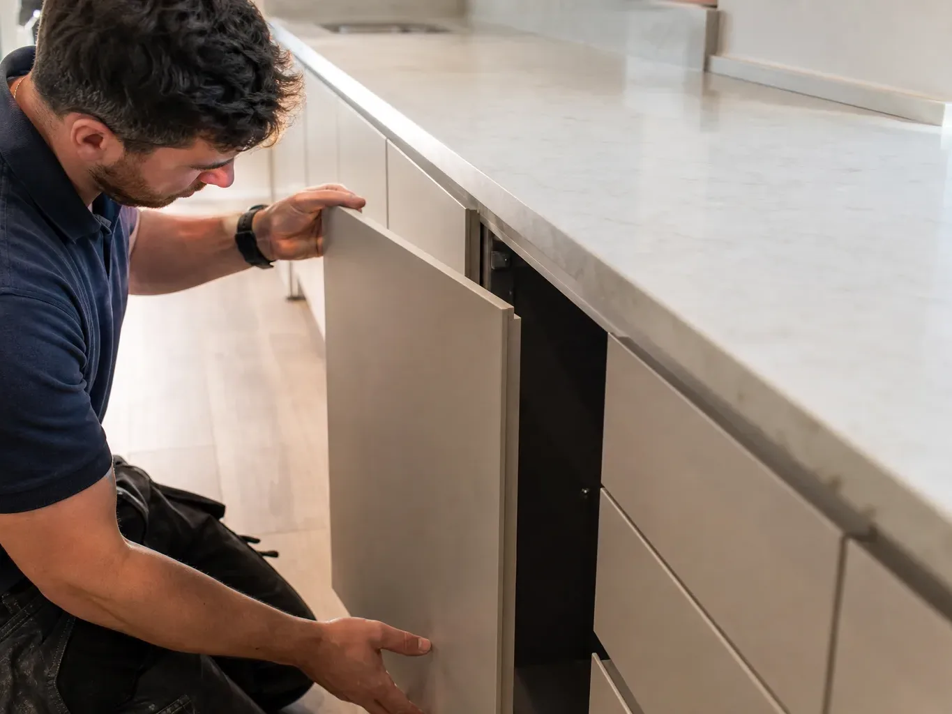 A kitchen fitter securing the final handleless cabinet door onto a base unit at the end of a long fitted run while a freshly installed quartz worktop with visible edge detail stretches across the top of the completed cabinets in a bright domestic kitchen