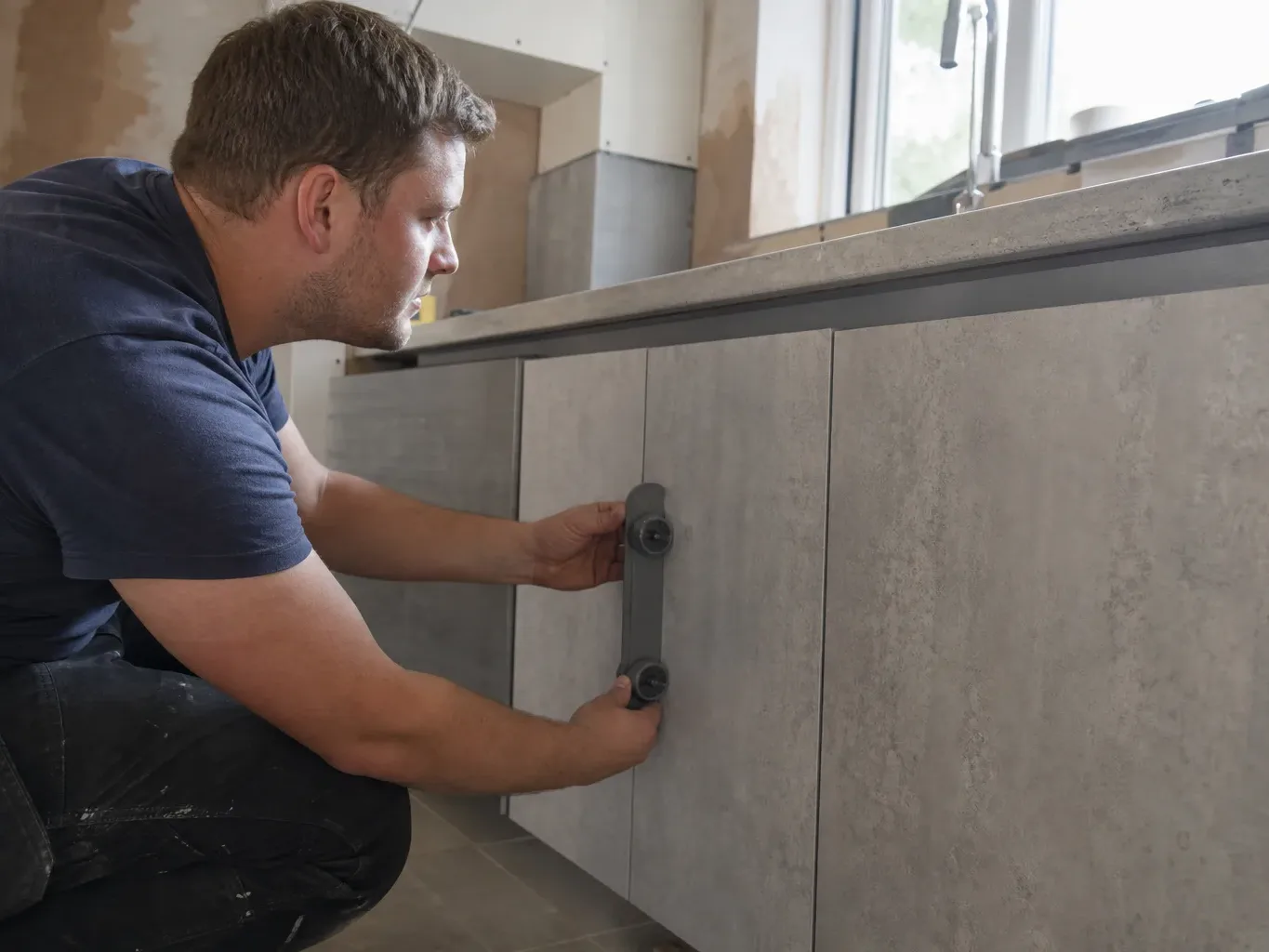 A kitchen fitter standing at a base unit run and pressing a cabinet alignment tool against the face of a handleless door to check it sits flush with the neighbouring panel in a mid-renovation domestic kitchen with natural light from a window above the sink